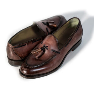 This loafer is crafted from premium leather featuring a hand-finished antique-effect patina in rich dark brown tones. The timeless design is enhanced by decorative tassels and hand-stitched detailing on the apron toe, making it a versatile choice for both formal and smart-casual looks. Expert craftsmanship and attention to detail ensure outstanding comfort, quality, and enduring style.